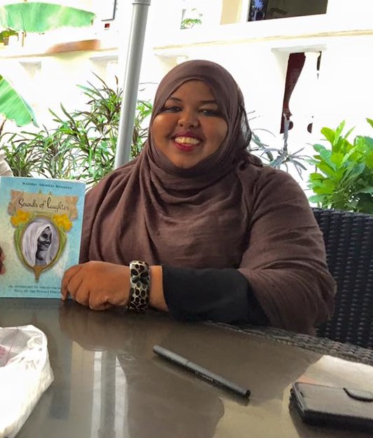 A woman smiling with a head cover sitting ona  desk and presenting a book 