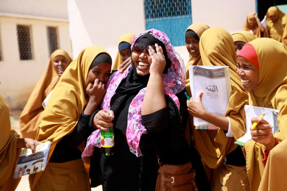 A joyful group of Somali schoolgirls in mustard-colored uniforms gather around a smiling woman in a pink floral headscarf, laughing and holding notebooks.