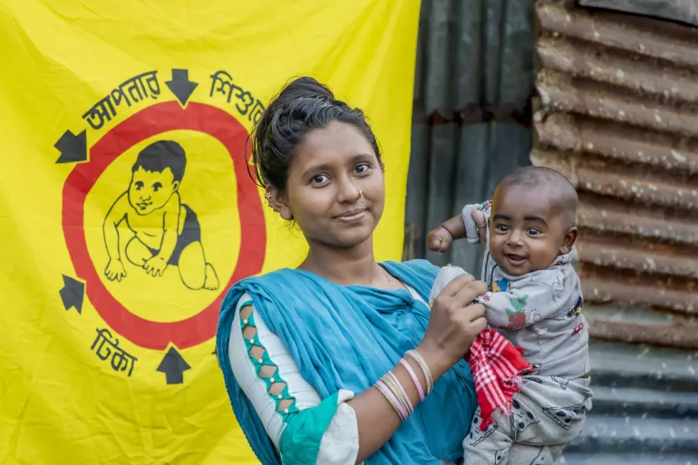 A young mother holds her smiling child beside a childhood immunisation awareness banner,